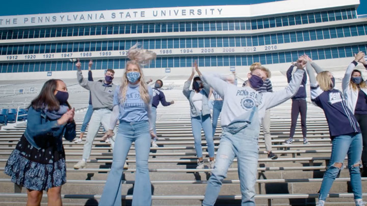 A group of a dozen musical theatre students dressed in Penn State apparel sing and dance in the stands of Beaver Stadium.